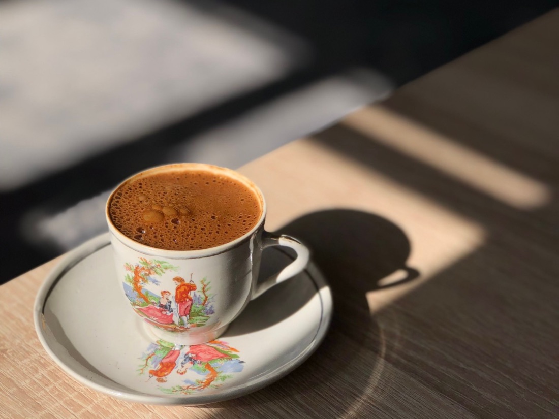 A cup of coffee on a patterned saucer in morning sunlight on a wooden table